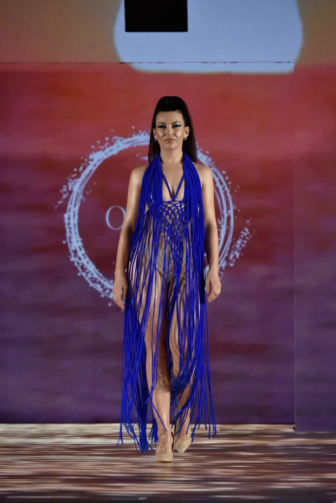 A model walks at New York Fashion Week runway wearing a floor-length olive green macramé net dress with long fringe tassels. The look features a high-neck halter design and is styled with bold teal eyeshadow and brown platform heels against a beach-themed backdrop.
